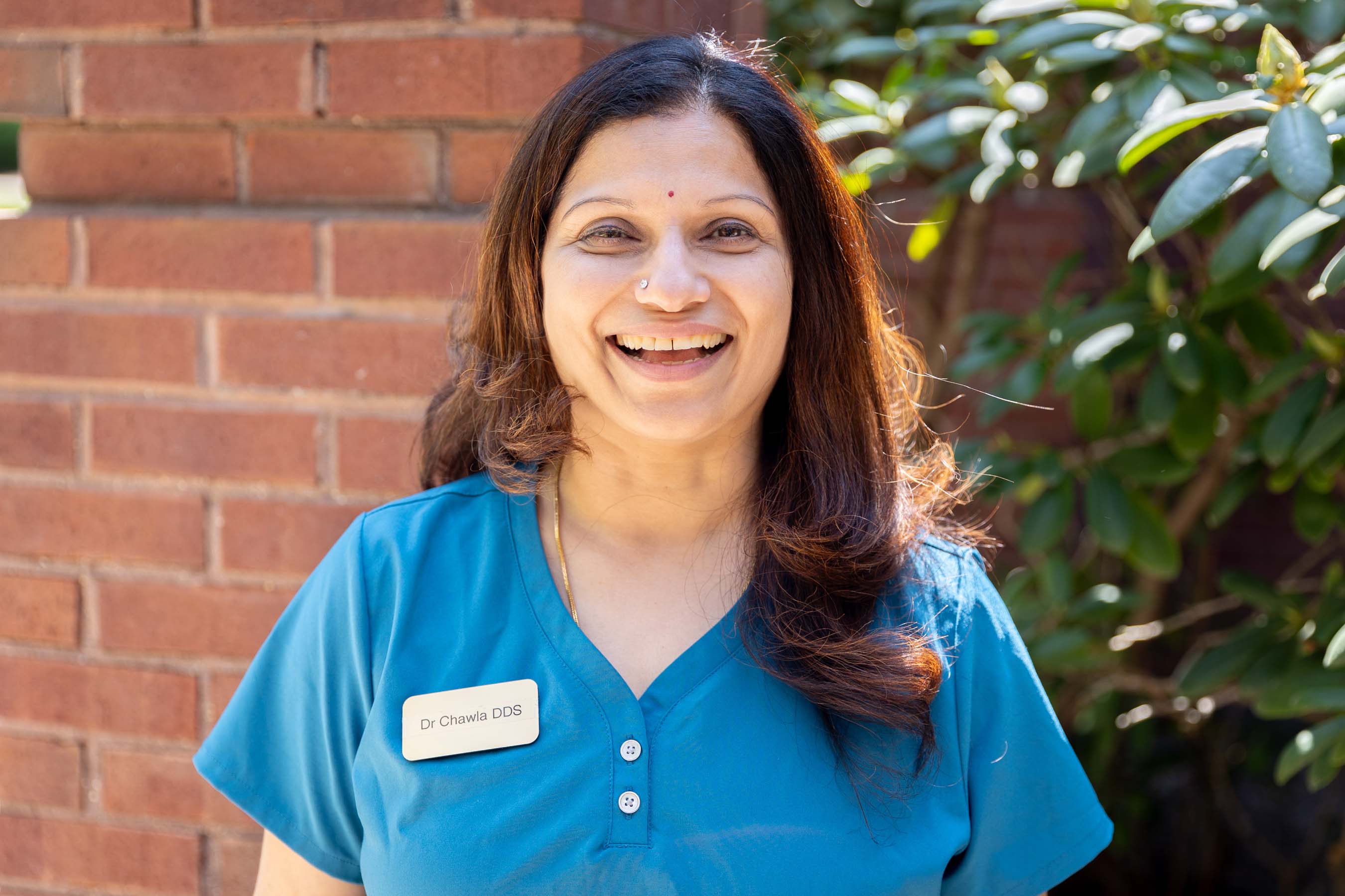 Smiling woman with brown hair in blue scrubs stands outdoors by a brick wall and green shrub.